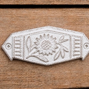 Puede incluir: Un tirador de cajón hexagonal de metal blanco con un diseño de girasol en relieve en el centro y patrones de líneas verticales en los lados. El herraje incluye dos tornillos de cabeza Phillips visibles para su montaje.