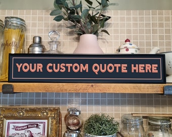 May include: Black chalkboard sign with white trim and the text "YOUR CUSTOM QUOTE HERE" in orange. The sign is hanging on a shelf above a kitchen counter with various kitchen items.