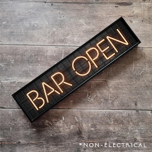 May include: A black framed sign with the words "BAR OPEN" in yellow neon lettering. The sign is on a woodgrain background.