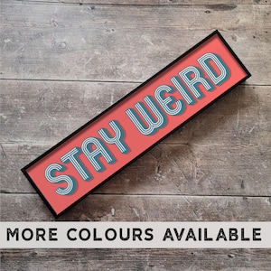 May include: A framed sign with a red background and black frame. The sign says "Stay Weird" in white and blue lettering.
