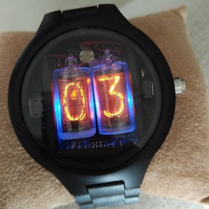 Könnte beinhalten: Eine schwarze Armbanduhr mit einem runden Zifferblatt und einem schwarzen Metallband. Das Zifferblatt der Uhr hat eine klare Glasabdeckung und zeigt die Zeit mit zwei orangefarbenen digitalen Röhren an. Die Röhren zeigen die Zahlen "03" an.