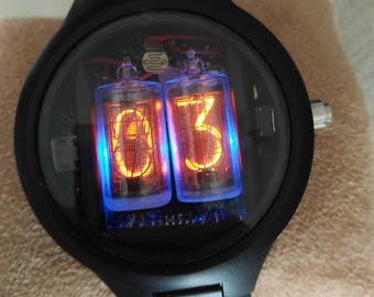 May include: A black wristwatch with a round face and a black metal band. The watch face has a clear glass cover and displays the time using two orange digital tubes. The tubes display the numbers "03".