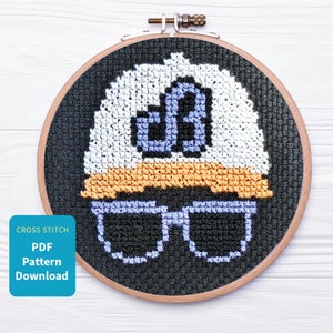 May include: Cross stitch pattern of a person wearing a white hat with a blue letter 'B' on it and black sunglasses. The pattern is in a wooden embroidery hoop.