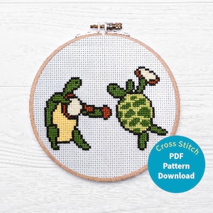 May include: Cross stitch pattern of two green turtles, one holding a flower and the other boxing. The pattern is in a round wooden embroidery hoop. The text "Cross Stitch PDF Pattern Download" is on a blue circle below the hoop.
