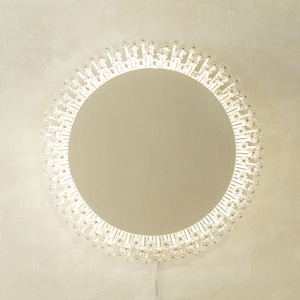 Op de afbeelding: Een ronde spiegel met een decoratieve, kristalachtige rand. Het oppervlak van de spiegel is lichtbeige en de rand bestaat uit vele kleine, heldere en sprankelende elementen. Een netsnoer loopt van onderen.