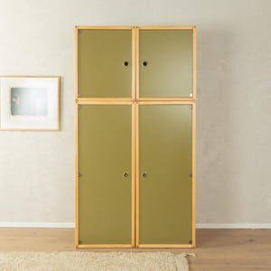 Puede incluir: Un armario de cuatro puertas con paneles verde oliva y un marco de madera clara. El armario está compuesto por cuatro secciones rectangulares apiladas. Una imagen enmarcada cuelga de la pared a la izquierda. Hay una alfombra en el suelo.