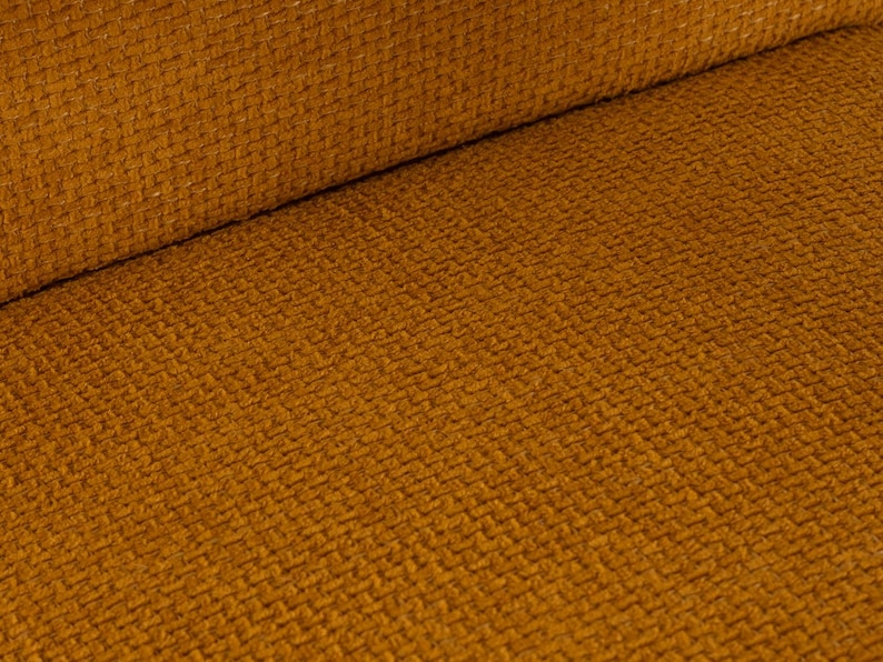 May include: Close-up of a textured fabric in a warm, golden-brown hue. The material has a woven appearance with a subtle diagonal pattern. The fabric is folded over, revealing a rolled edge.