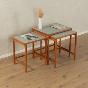 May include: Three wooden nesting tables with light grey tops. The tables are arranged in a staggered formation, with the largest table at the back and the smallest table at the front. The tables are made of a light brown wood and have a simple, modern design.