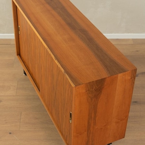 May include: A mid-century modern wooden cabinet with a rectangular shape. The cabinet has a warm brown finish with darker wood grain accents. It features a sliding door and sits on short, black metal legs. The cabinet is designed for storage.