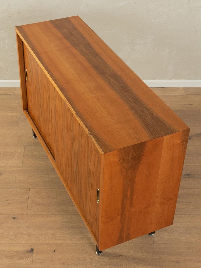 May include: A mid-century modern wooden cabinet with a rectangular shape. The cabinet has a warm brown finish with darker wood grain accents. It features a sliding door and sits on short, black metal legs. The cabinet is designed for storage.