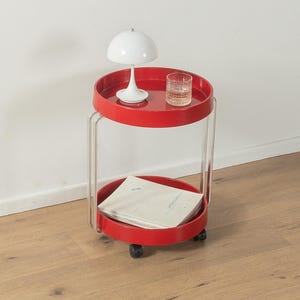 May include: A red, two-tiered rolling side table with a white lamp and a glass of water on the top tier. The bottom tier holds a stack of papers. The table has clear acrylic supports and black wheels.