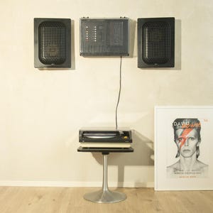 May include: A retro stereo system featuring two black speakers, a control panel, and a turntable on a silver stand. A framed David Bowie poster leans against the wall. The setup is on a light wood floor.