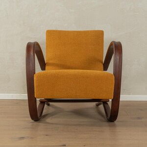 May include: An orange upholstered armchair with dark wooden curved arms and legs. The chair has a rectangular back and seat cushion. The wooden frame has a curved design. The chair is set against a neutral wall and wooden floor.