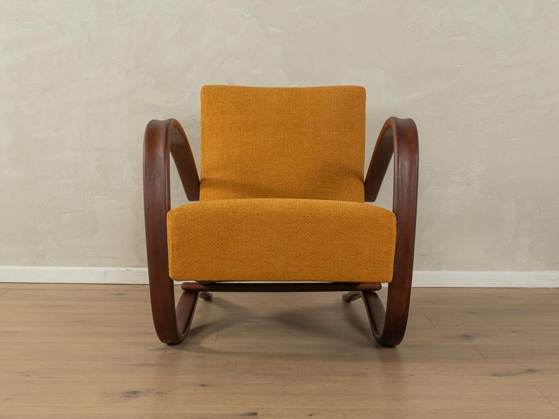 May include: An orange upholstered armchair with dark wooden curved arms and legs. The chair has a rectangular back and seat cushion. The wooden frame has a curved design. The chair is set against a neutral wall and wooden floor.