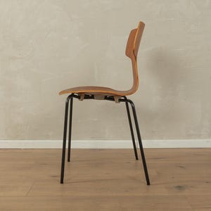 Puede incluir: Una silla de madera vintage con respaldo y asiento curvos. La silla tiene un asiento y respaldo de madera marr&oacute;n claro, con patas de metal negro. La silla se muestra de perfil sobre un fondo neutro.