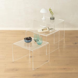 Peut inclure: Un ensemble de trois tables gigognes en acrylique transparent de hauteurs variables. Les tables présentent un design lisse en forme de U avec des coins arrondis. Les surfaces supportent une lampe blanche, un vase en verre et deux verres.