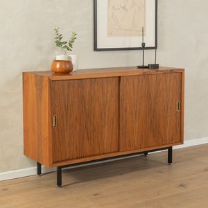 May include: A mid-century modern wooden cabinet with sliding doors. The cabinet is made of a warm-toned wood with a visible grain pattern. It has brass handles and sits on black metal legs. A small vase with greenery and a framed artwork are on top.