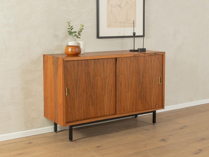 May include: A mid-century modern wooden cabinet with sliding doors. The cabinet is made of a warm-toned wood with a visible grain pattern. It has brass handles and sits on black metal legs. A small vase with greenery and a framed artwork are on top.