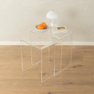 Op de afbeelding: Transparante acryl bijzettafel met een nestontwerp. De tafel is bekroond met een witte lamp, een kom met sinaasappels en een boek. De tafel staat op een hardhouten vloer.