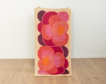 May include: A rectangular, vintage wall hanging with a beige background and a vibrant floral design. The flowers are composed of layered circles in shades of red, pink, and orange. The piece is displayed against a white wall and wooden floor.