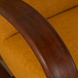 May include: Close-up of a mid-century modern armchair. The curved armrest is made of dark brown wood, contrasting with the yellow-orange textured fabric of the seat and backrest. The design suggests a focus on comfort and style.