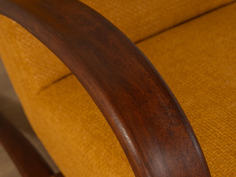 May include: Close-up of a mid-century modern armchair. The curved armrest is made of dark brown wood, contrasting with the yellow-orange textured fabric of the seat and backrest. The design suggests a focus on comfort and style.