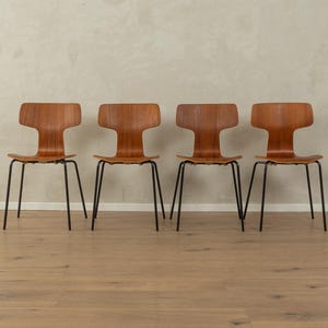 Puede incluir: Cuatro sillas de estilo moderno de mediados de siglo con un respaldo distintivo en forma de T. Las sillas presentan un asiento y respaldo de madera marr&oacute;n c&aacute;lido, contrastados por elegantes patas de metal negro. Las sillas est&aacute;n dispuestas en fila.