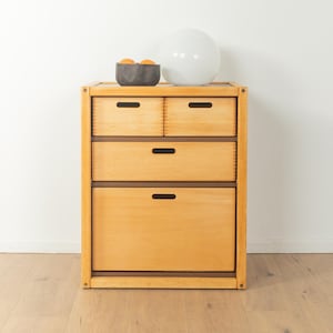 Puede incluir: Un mueble de madera clara con cuatro cajones. La parte superior del mueble tiene un cuenco gris oscuro con tres naranjas y una esfera blanca. Los cajones tienen detalles en marrón oscuro y tiradores recortados. El mueble está sobre un suelo de madera clara.