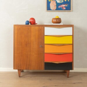 Op de afbeelding: Een houten kast in het midden van de eeuw met een deur en vijf laden in wit, geel, oranje, rood en zwart. Decoratieve items staan bovenop, waaronder een blauwe bol en een rode sculptuur. Boven hangt een kleurrijk schilderij.