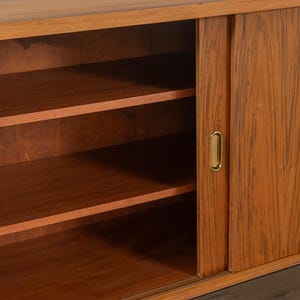 May include: A mid-century modern wooden cabinet with sliding doors and interior shelves. The cabinet is made of a warm-toned wood with a visible grain pattern. The sliding door has a brass-colored handle.