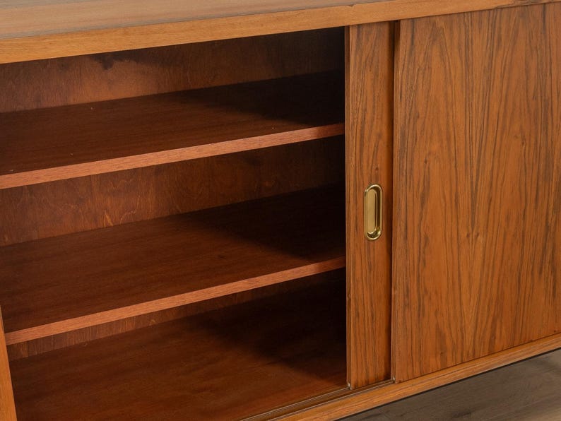 May include: A mid-century modern wooden cabinet with sliding doors and interior shelves. The cabinet is made of a warm-toned wood with a visible grain pattern. The sliding door has a brass-colored handle.