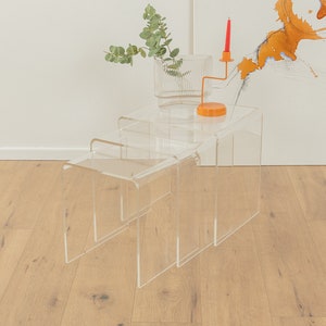 Op de afbeelding: Drie in elkaar passende tafels van helder acryl met een rode kaarsenhouder en een kleine oranje basis op de bovenste tafel. De tafels staan op een lichte houten vloer.
