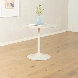 Könnte beinhalten: Ein weißer, runder Beistelltisch mit einem einzelnen Sockel. Eine hellblaue Vase mit Grün steht auf dem Tisch, zusammen mit einem Stapel weißer Papiere. Der Tisch steht auf einem hellen Holzboden.