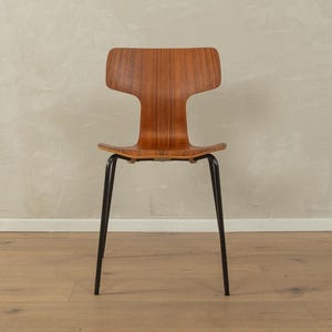 Puede incluir: Silla de estilo moderno de mediados de siglo con asiento y respaldo de madera curvados. La silla tiene un respaldo distintivo en forma de T y est&aacute; sostenida por cuatro patas de metal negro. La madera tiene un tono marr&oacute;n c&aacute;lido y la silla est&aacute; sobre un fondo neutro.