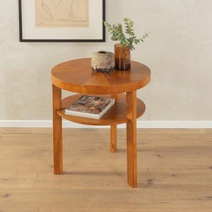 Può includere: Un tavolino rotondo in legno con due ripiani. Il tavolo ha una finitura in legno naturale ed è decorato con un motivo geometrico. Il tavolo è posto su un pavimento in legno e ha un libro e due vasi sul ripiano superiore.