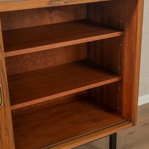 May include: A mid-century modern wooden cabinet with three shelves. The cabinet is made of a warm-toned wood with a visible grain. It has a sliding door and black metal legs. The shelves are empty.