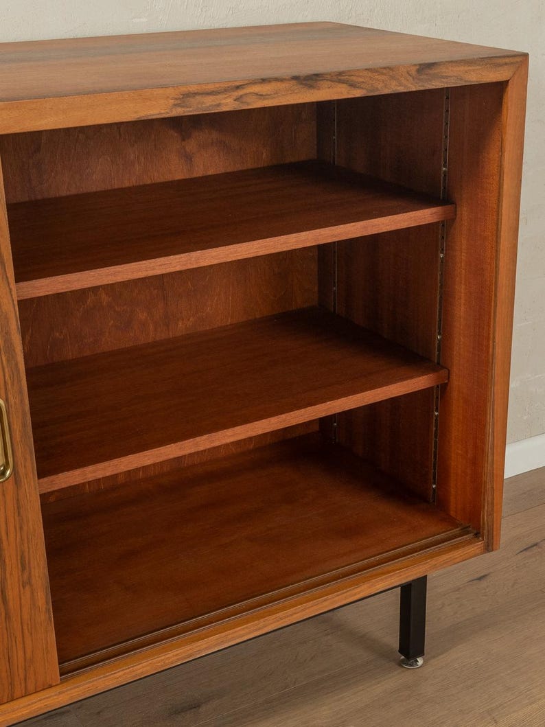 May include: A mid-century modern wooden cabinet with three shelves. The cabinet is made of a warm-toned wood with a visible grain. It has a sliding door and black metal legs. The shelves are empty.