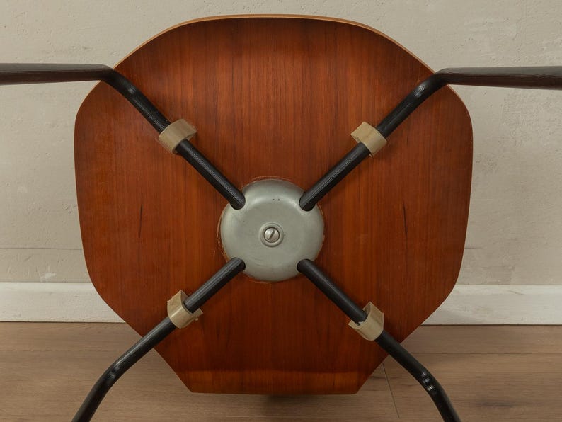 Puede incluir: Vista inferior de una silla moderna de mediados de siglo. La silla presenta un asiento de madera, patas de metal negro y un soporte central de metal. Las patas est&aacute;n unidas al asiento con conectores de goma o pl&aacute;stico de color beige.