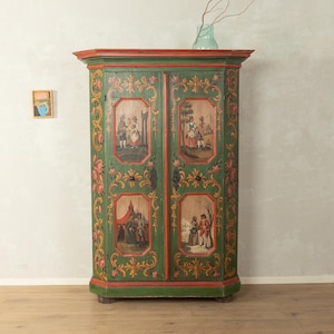 Op de afbeelding: Een hoge, groene houten kast met rode randen aan de boven- en onderkant. De twee deuren zijn voorzien van handgeschilderde florale krulmotieven in rood en goud, en vier rechthoekige panelen met afbeeldingen van figuren in een buitenomgeving.
