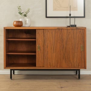 May include: A mid-century modern wooden cabinet with a sliding door and open shelving. The cabinet is made of a warm-toned wood with a visible grain. It has brass hardware and sits on black metal legs. Decorative items are on top.