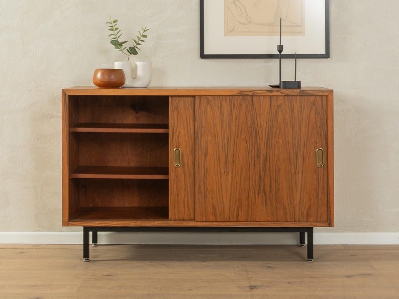 May include: A mid-century modern wooden cabinet with a sliding door and open shelving. The cabinet is made of a warm-toned wood with a visible grain. It has brass hardware and sits on black metal legs. Decorative items are on top.