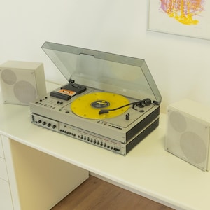 Peut inclure: Une chaîne stéréo vintage avec une platine, un lecteur de cassettes et deux enceintes. La platine a un disque jaune dessus. Le système est argenté et noir, et les enceintes sont blanches. Le système est sur un bureau blanc.