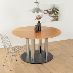 Könnte beinhalten: Ein runder Holztisch mit einem verchromten Untergestell und vier Beinen. Ein Stuhl aus verchromtem Draht sitzt neben dem Tisch. Eine Trockenblumen-Arrangement steht auf dem Tisch.