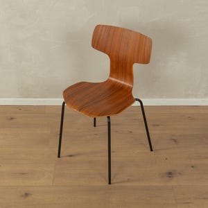 Puede incluir: Una silla moderna de mediados de siglo con asiento y respaldo curvos de madera. La silla tiene un dise&ntilde;o distintivo recortado en la parte superior del respaldo y se apoya en cuatro patas delgadas de metal negro. La veta de la madera es visible.