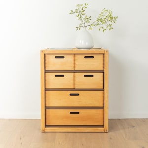 Puede incluir: Un aparador de madera clara con cinco cajones. Los cajones tienen tiradores marrones oscuros y están dispuestos en una configuración 2-2-1. Un jarrón blanco con ramas verdes se encuentra encima del aparador.