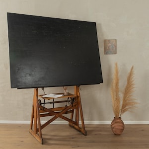 Può includere: Un tavolo da disegno vintage con una superficie di disegno rettangolare nera e una cornice in legno. Il tavolo è su un pavimento in legno marrone chiaro.