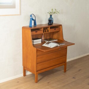 Puede incluir: Un escritorio secretario de madera con un acabado marrón medio cálido. El escritorio cuenta con una superficie de escritura abatible, cajones y pequeños compartimentos. Un reloj azul y un jarrón con vegetación encima. El escritorio mide unos 102 cm de alto.