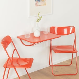 Könnte beinhalten: Ein leuchtend orangefarbener, an der Wand montierter Klapptisch mit zwei passenden orangefarbenen Klappstühlen. Auf der Tischplatte stehen zwei klare Glasbecher und eine weiße Keramikvase mit einem kleinen grünen Blattzweig.