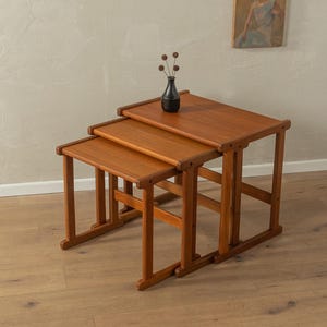 Può includere: Tre tavolini in legno nidificati di colore marrone chiaro. I tavolini sono impilati uno sopra l'altro e hanno un design semplice e moderno.