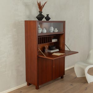 Peut inclure: Un meuble en bois de style milieu du siècle avec un bureau rabattable, une partie supérieure vitrée et des compartiments de rangement. Le meuble est de couleur marron chaud et une petite chaise blanche est à droite. Des vases décoratifs sont posés dessus.